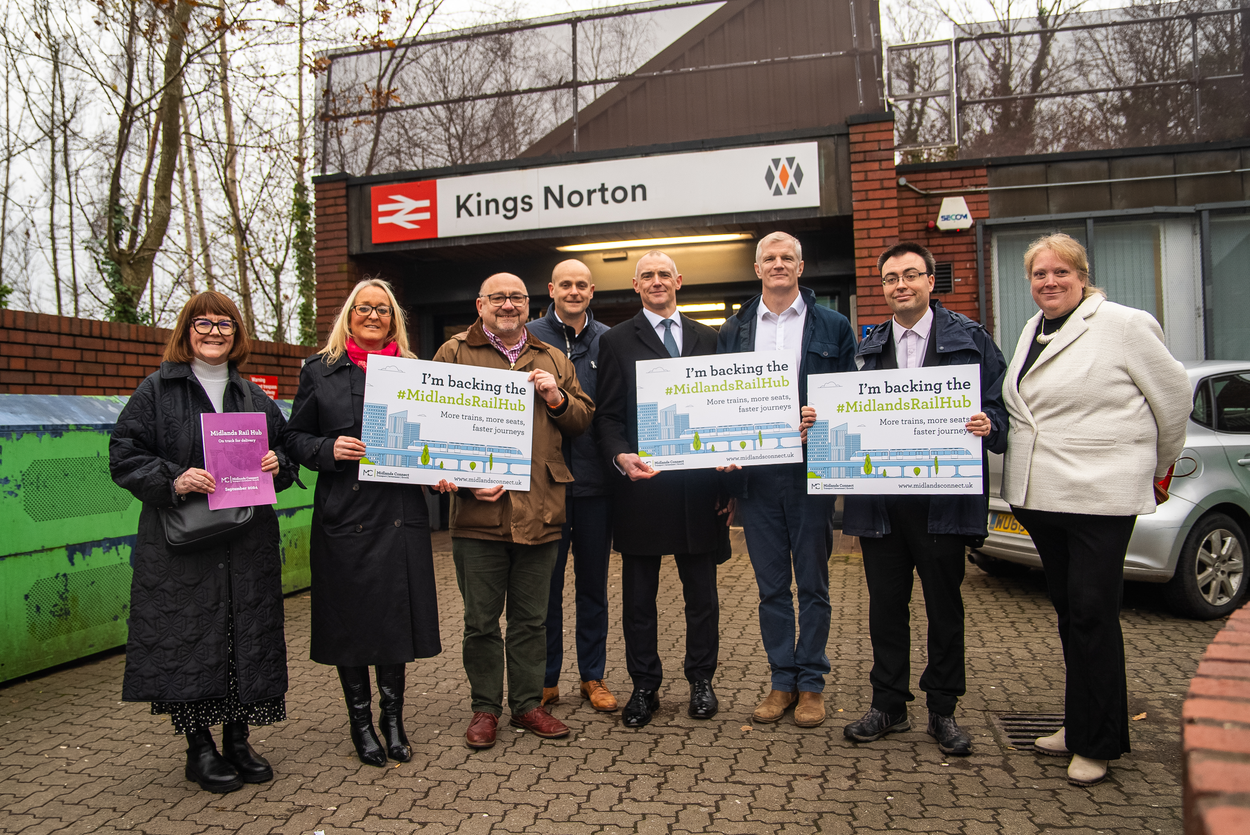 Eight people stood at Kings Norton railway station. Three are holding placards which read "I'm backing the Midlands Rail Hub"